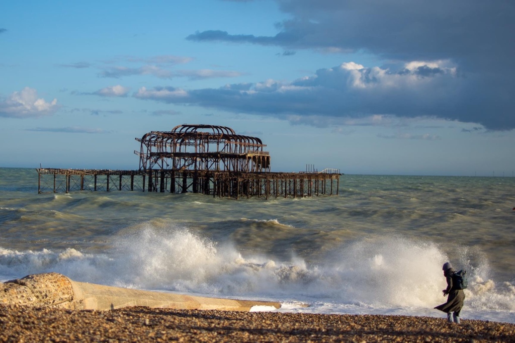 An image of the ruined skeletal remains of the pier in a rough sea. Surf is breaking onto a pebbly beach. In the bottom right hand corner a person stands, their clothes being whipped up by the wind. The sky above is mostly blue with some grey clouds. 