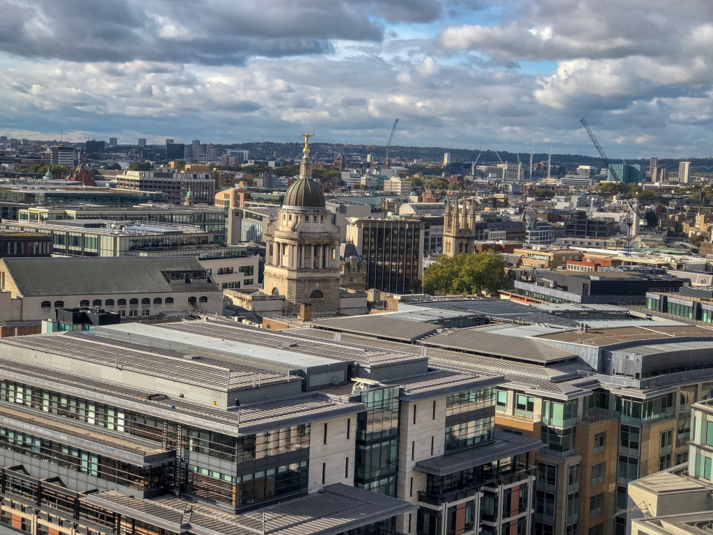 View of London from high up. In the cetnre is a dome with a gold statue of Justice with scales. There are many flat rooves in the foreground and amongst them are trees. In the distance there are some cranes and hills. The sky is cloudy with patches of blue. 
